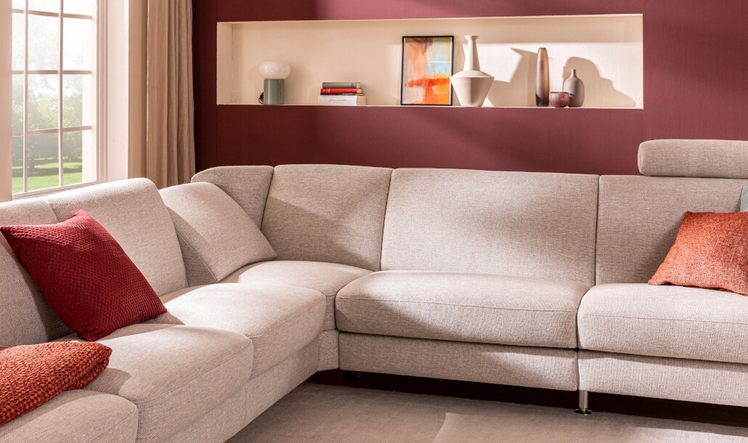 Modernes Wohnzimmer mit hellgrauem Ecksofa Canto, roten Kissen und Dekoartikeln auf einem burgunderfarbenen Wandregal. Großes Fenster links.