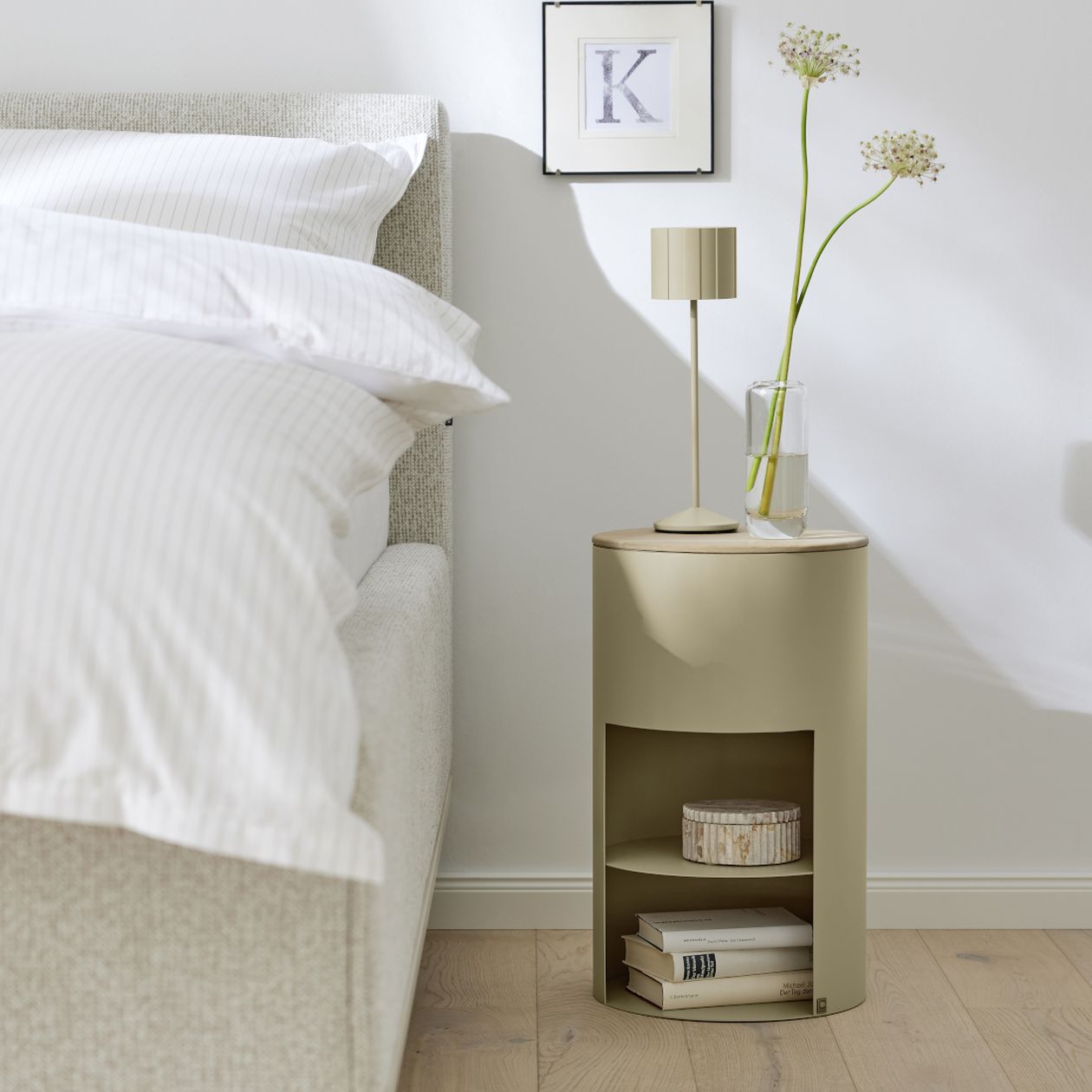 Minimalistisches Schlafzimmer mit beigem Nachttisch und Lampe, weißer Bettwäsche, Topfpflanzen und einem gerahmten Kunstwerk mit dem Buchstaben „K“ an einer hellen Wand.