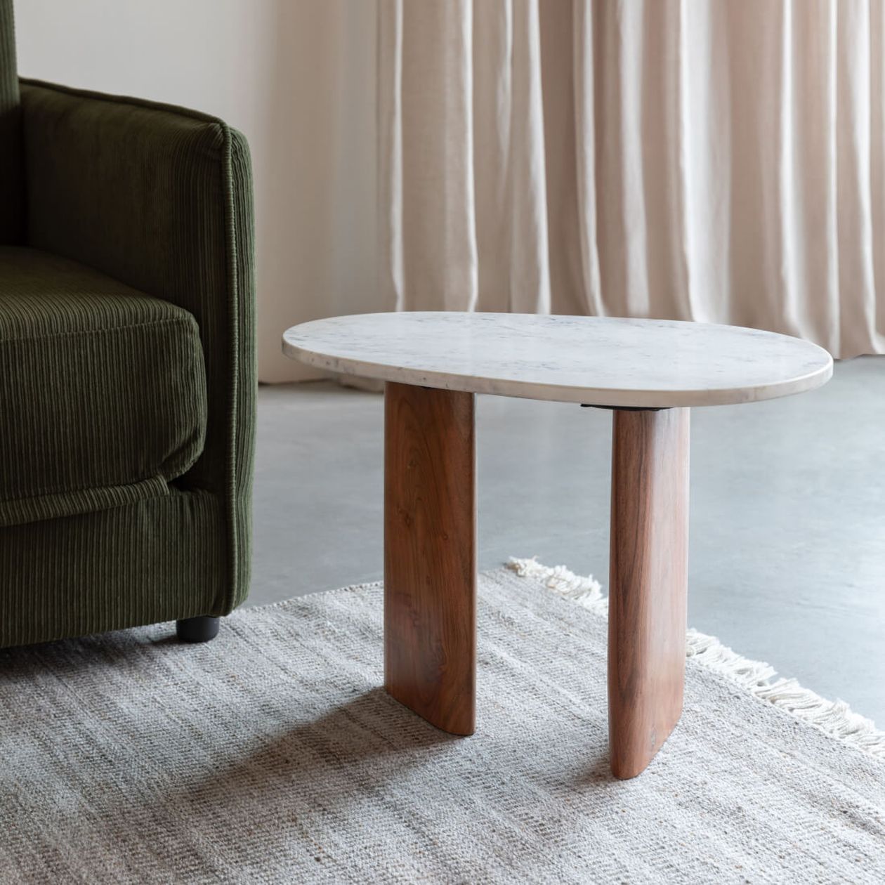 Ovaler Couchtisch mit Marmorplatte und Holzbeinen auf einem gewebten Teppich neben einem grünen Cordsofa und beigen Vorhängen.