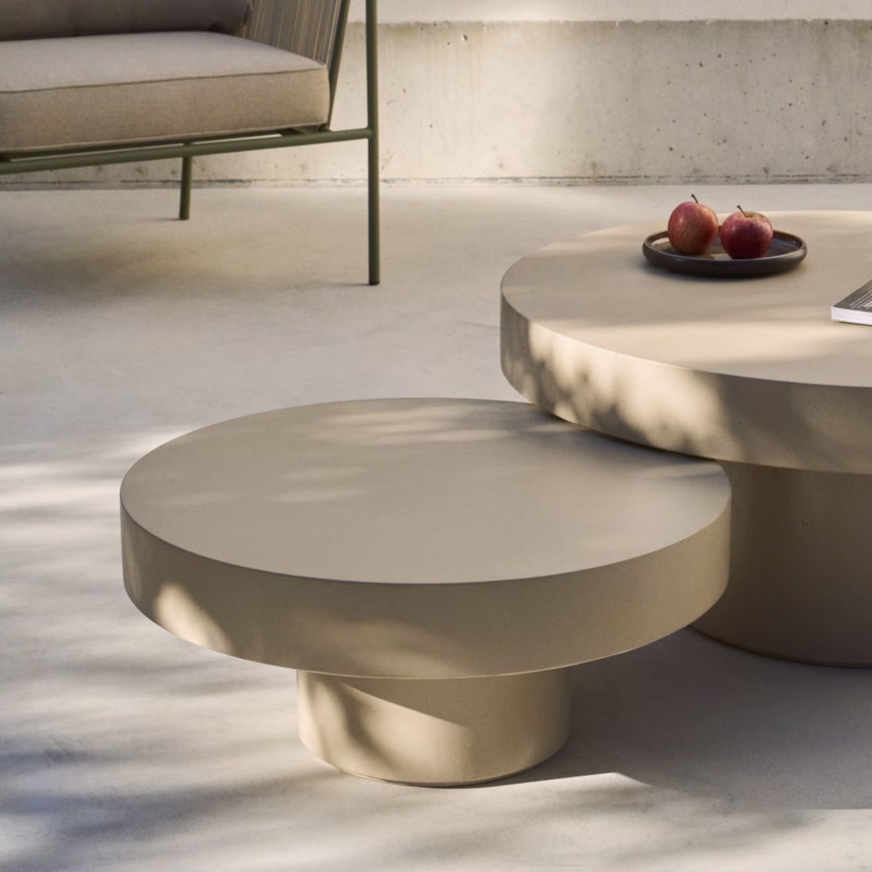 Moderne beigefarbene runde Tische auf einer sonnigen Terrasse, mit Äpfeln auf einem Teller und einem Buch. Im Hintergrund steht ein Stuhl.