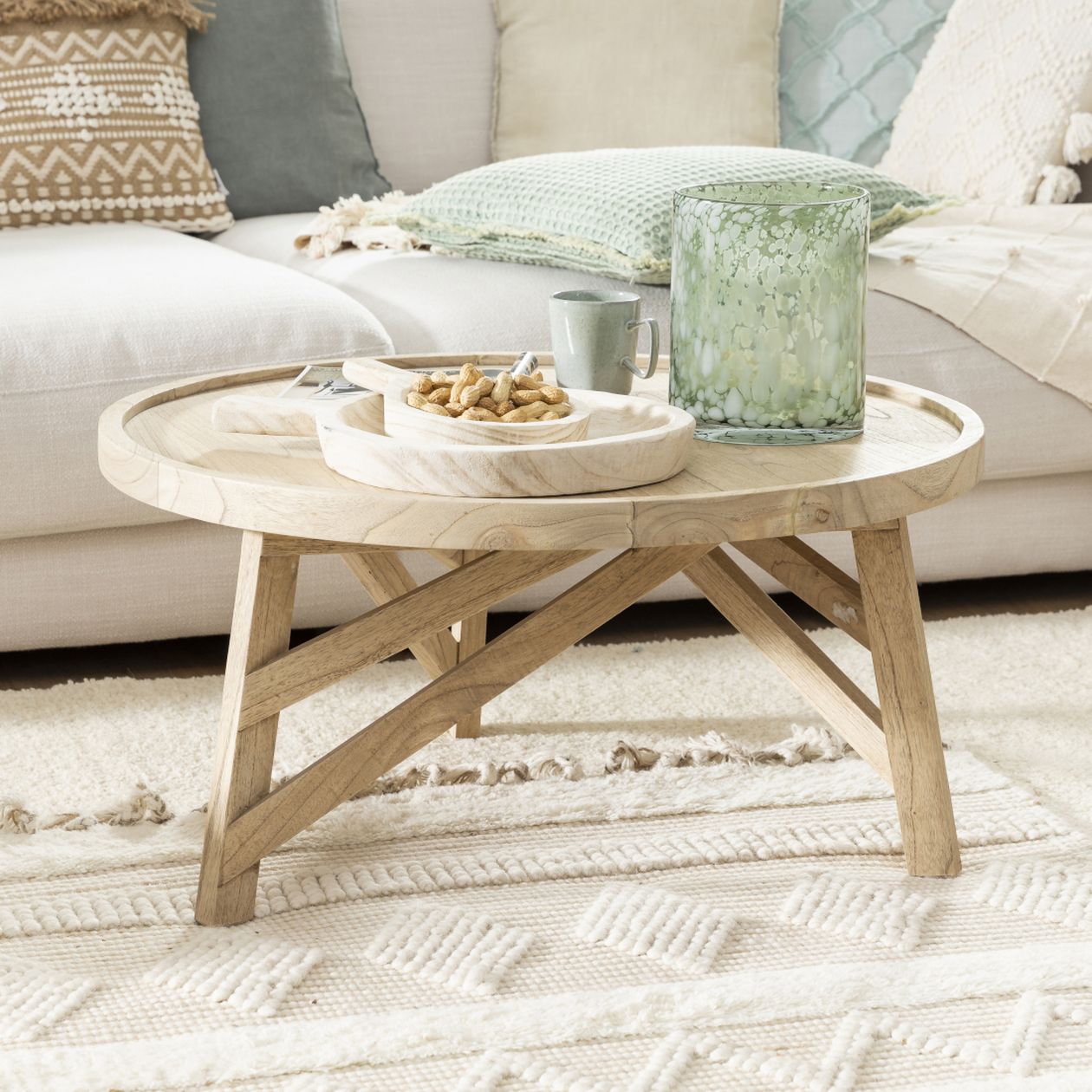 Holz-Couchtisch mit Snacks und einem grünen Kerzenhalter in einem gemütlichen Wohnzimmer. Beigefarbenes Sofa und strukturierte Kissen im Hintergrund.