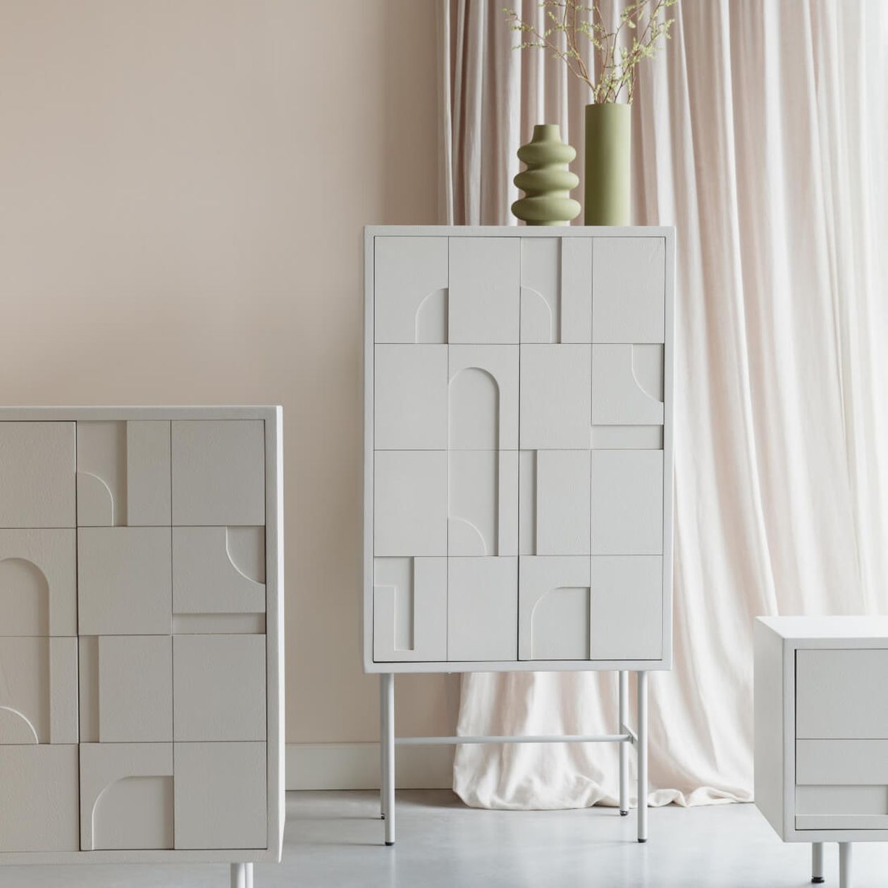 Hellgraues Highboard mit Türen, die mit geometrischen Einschnitten versehen sind. Darüber zwei grüne Vasen.