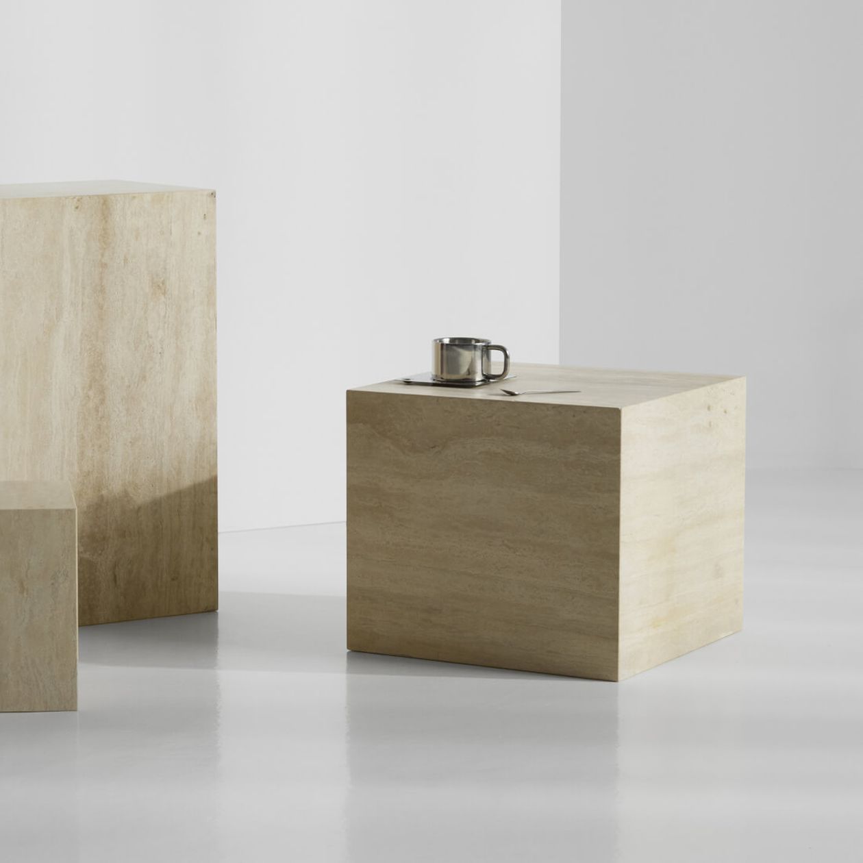Minimalistischer weißer Raum mit drei beigen Steinwürfeln; auf dem nächstgelegenen Würfel stehen ein Edelstahlbecher und ein Löffel.