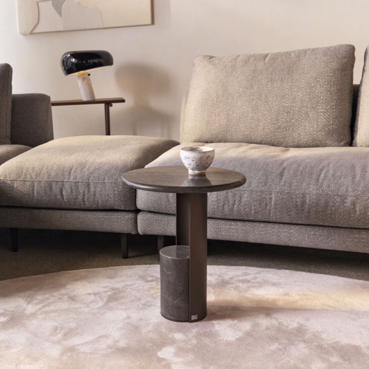 Beiges Ecksofa mit einem kleinen dunklen runden Beistelltisch, auf dem eine Keramiktasse auf einem hellen Teppich steht; moderne Lampe und Beistelltisch im Hintergrund.