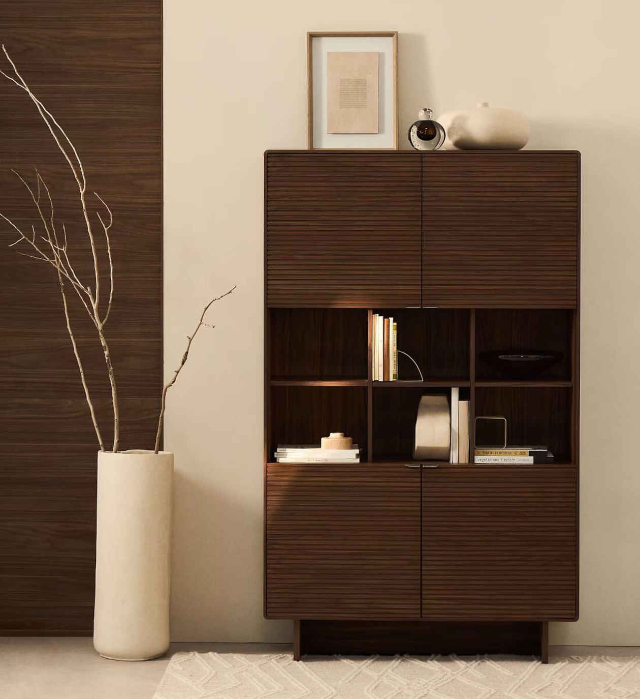 Holzschrank mit offenen Regalen, in denen Dekoartikel ausgestellt sind, flankiert von einer hohen weißen Vase mit Zweigen. Wandkunst und Keramik schmücken die Oberseite.