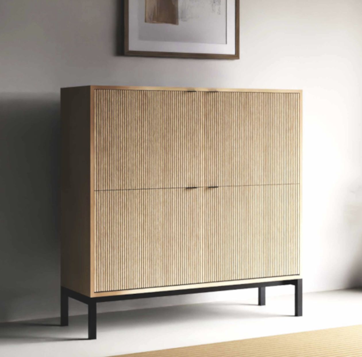 Heller Holzschrank mit vertikalen Lamellen vor einer grauen Wand, darauf eine weiße Lampe, eine große weiße Vase und eine grüne Vase mit Trockenblumen.