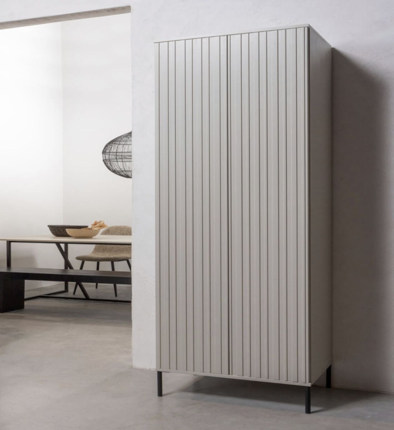 Ein hoher, schmaler Schrank mit vertikalen Rillen steht in einem minimalistischen Raum mit einer Bank, einer Schale und einer Drahtgitter-Leuchte im Hintergrund.