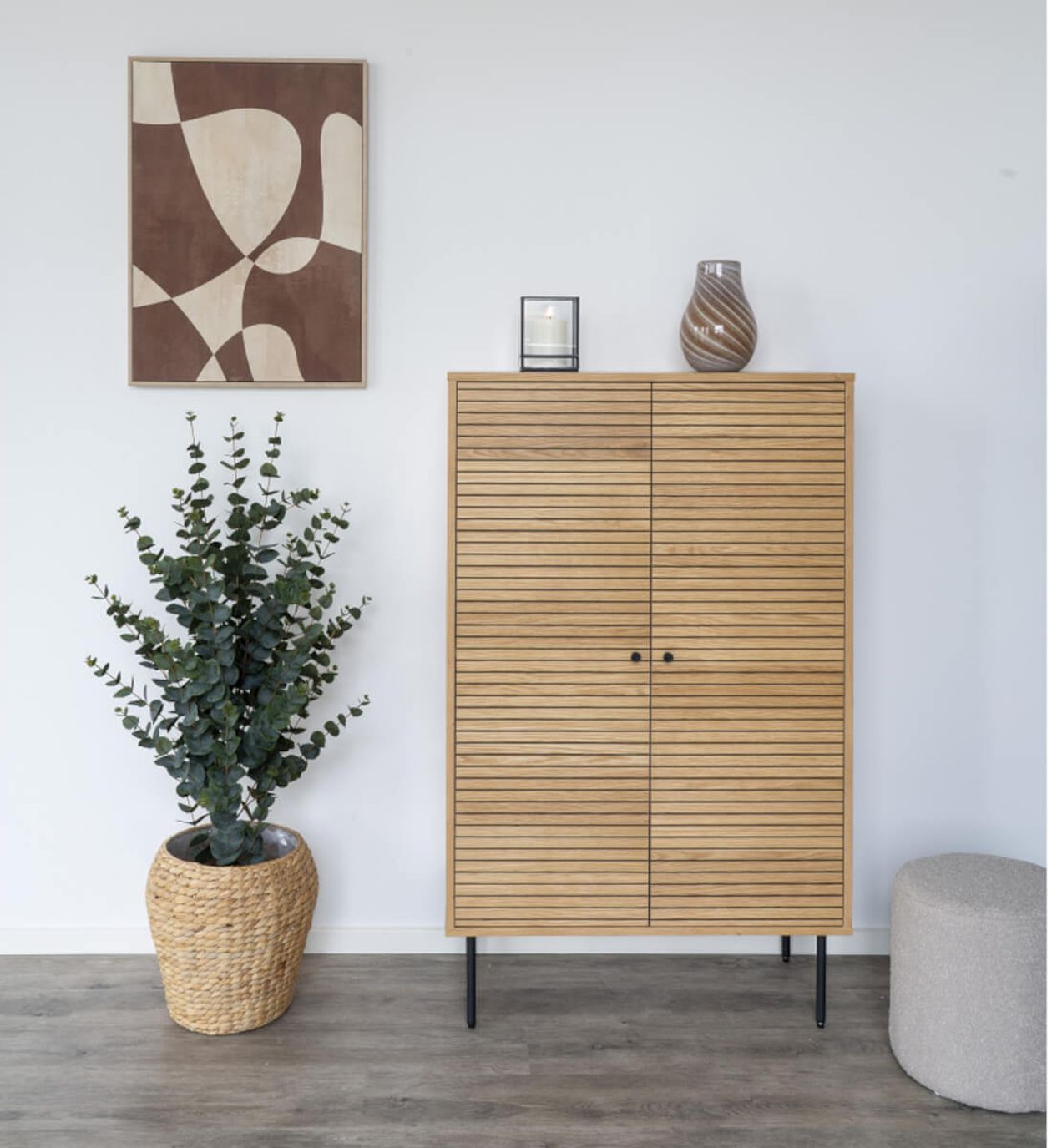 Ein Holzschrank mit Streifenmuster steht neben einer Topfpflanze. Darauf stehen eine Vase und eine Kerze. An der weißen Wand hängt ein abstraktes Kunstwerk.