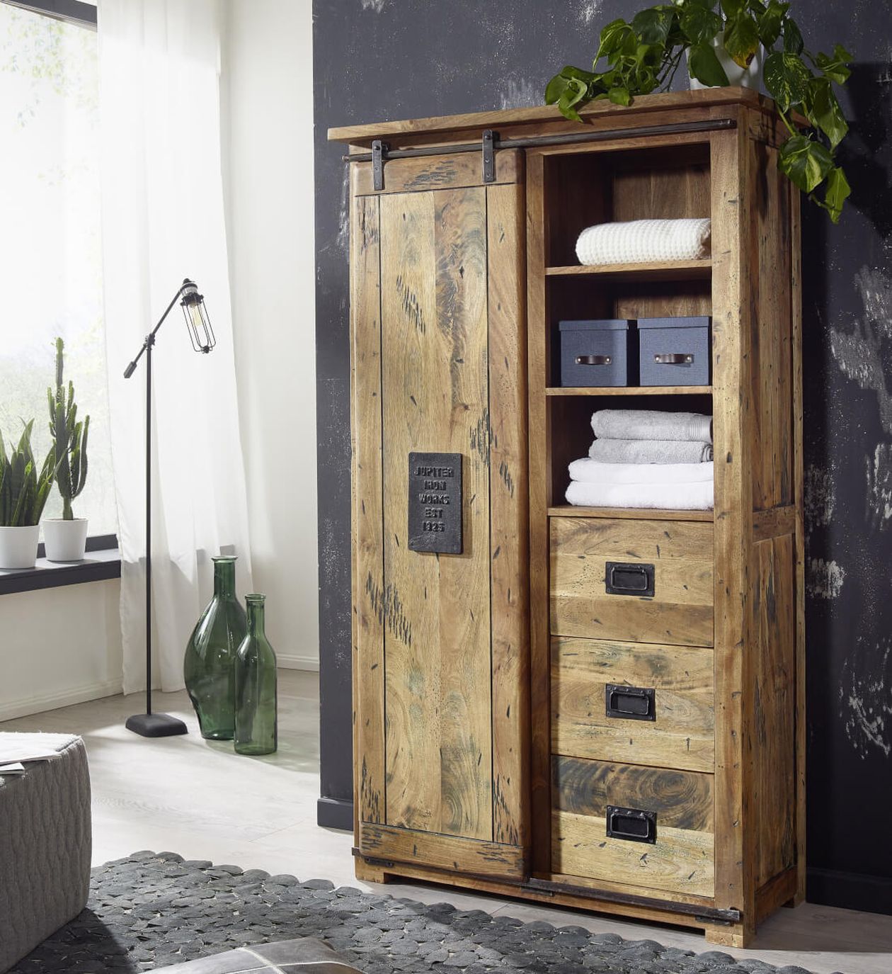 Rustikaler Holzschrank mit Schiebetür, offenen Regalen mit Handtüchern und Kisten, Schubladen darunter, neben einem Fenster mit Pflanzen und einer schwarzen Stehlampe.