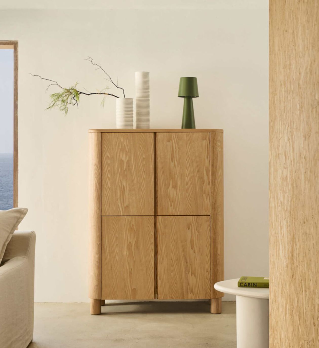 Holzschrank mit vier Türen, verziert mit gestapelten weißen Vasen, einer grünen Lampe und einem Ast. Beigefarbene Couch und Tisch in einem Zimmer mit Meerblick.