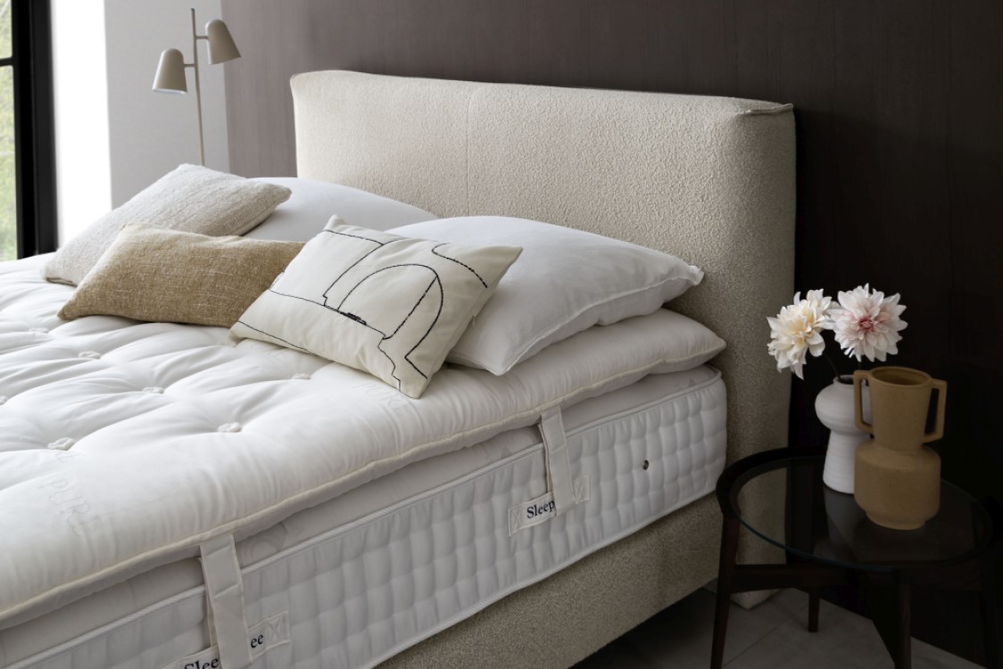 Ein ordentlich gemachtes Bett mit weißer Bettwäsche, beigen und weißen Kissen, neben einem dunklen Beistelltisch mit Blumen und dekorativen Vasen.