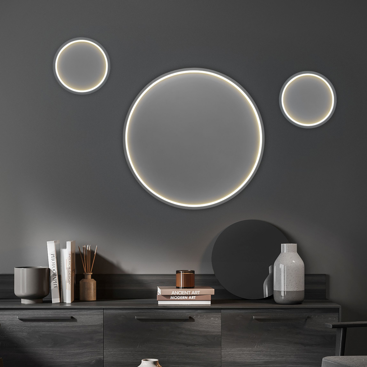 Die Wandleuchte Loop von Hell mit einem LED-Ring um eine graue Fläche hängt in drei Ausführungen an einer dunklen Wand über einer eleganten schwarzen Kommode.