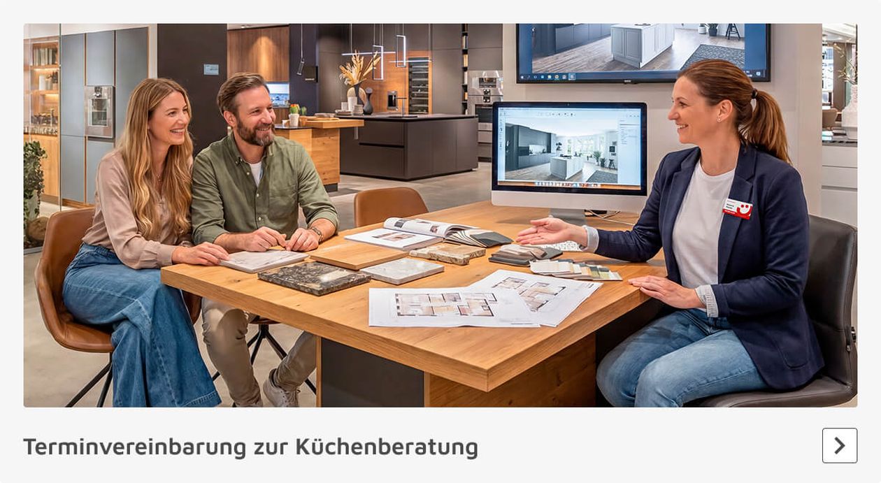 Drei Personen sitzen an einem Tisch in einem Küchenstudio und besprechen Küchenentwürfe anhand von Zeichnungen und Mustern auf dem Tisch.