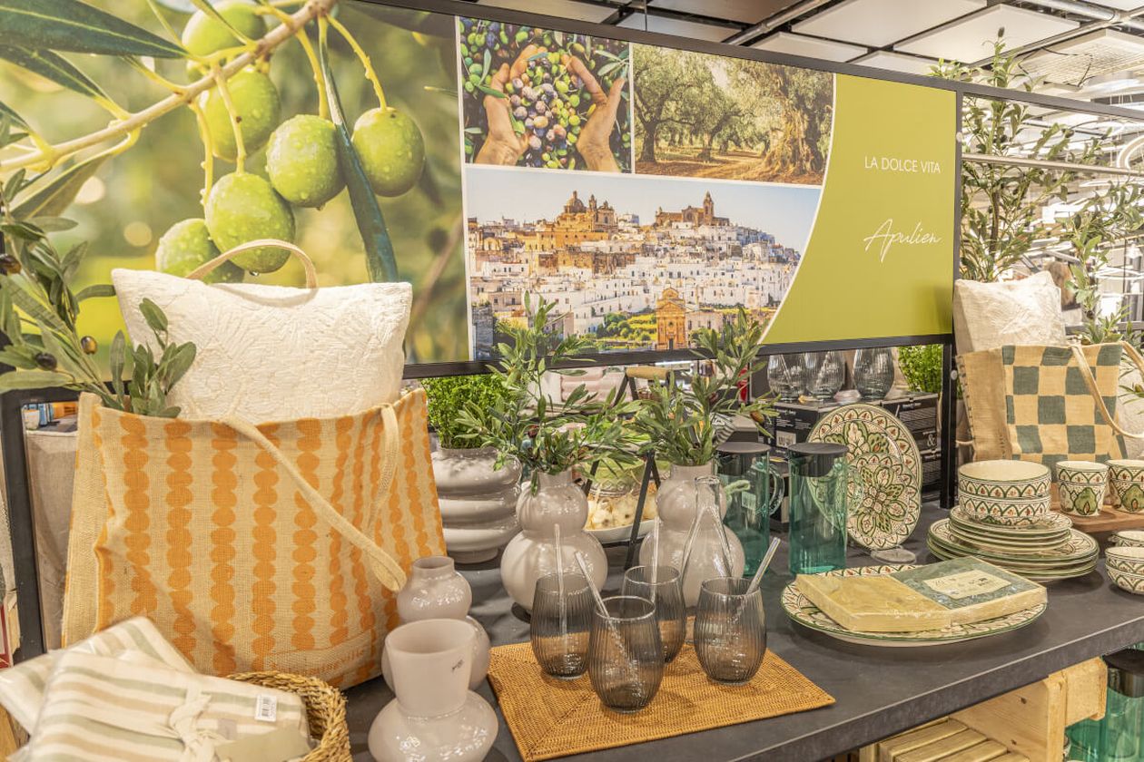 Ausstellung Wohnaccessoires im mediterranen Stil mit Keramik, Glaswaren und Textilien. Im Hintergrund sind Olivenbäume und Landschaften Süditaliens zu sehen.