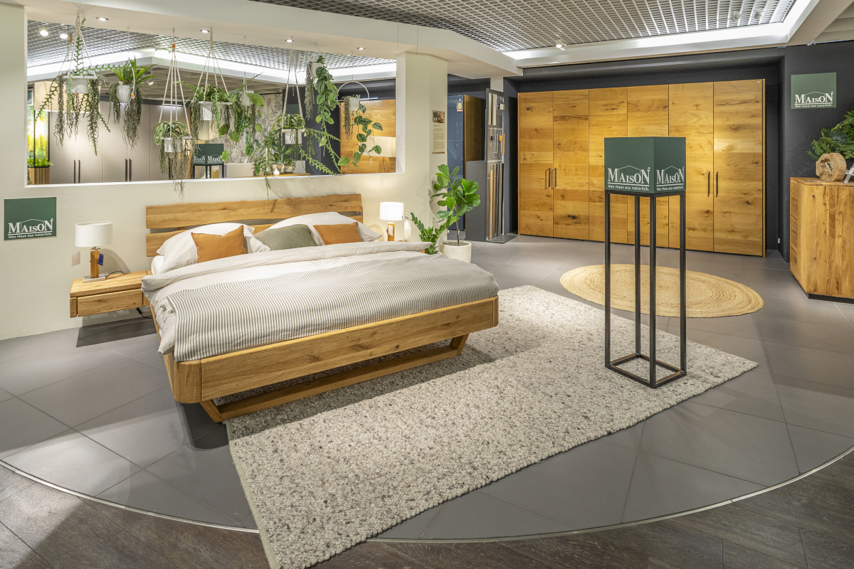 In einem Ausstellungsraum für Möbel der Marke MAISON steht das Massivholzbett Omaha mit integrierten Nachttischen und Füßen in Kufenform. Im Hintergrund befindet sich der sechstürige Kleiderschrank Miami aus Holz.