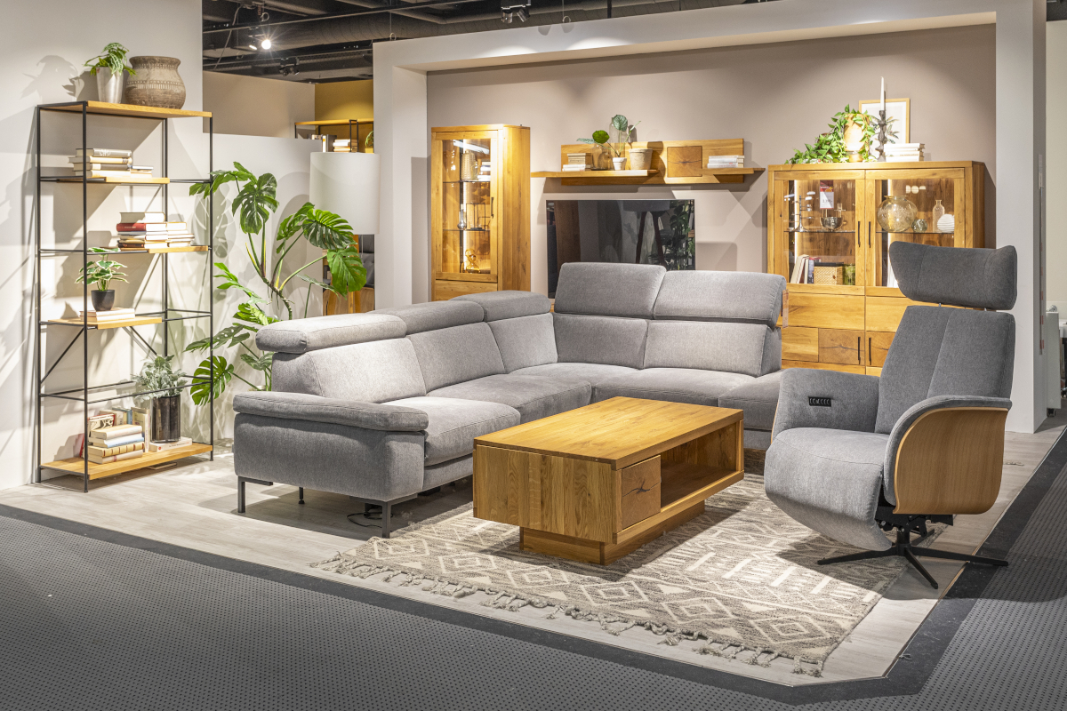 In einem Ausstellungsraum für Wohnzimmermöbel der Marke MAISON stehen der graue Sessel Cleveland mit Seitenverkleidung aus Holz, das graue Ecksofa Tampa und der Couchtisch Louisville aus Holz.