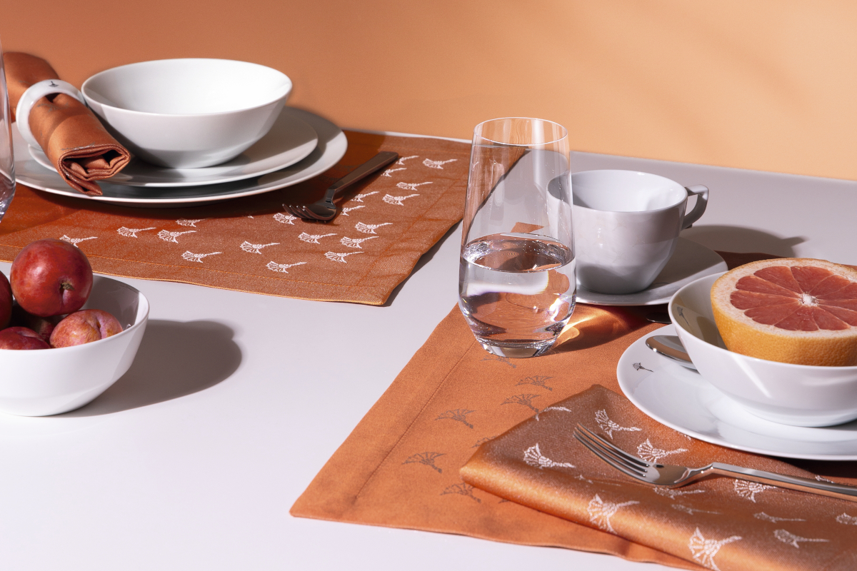 Gedeckter Tisch vor orangener Wand mit weißer Tischdecke und Servietten der Serie Faded in Apricot mit dem für JOOP! typischen Cornflower Muster