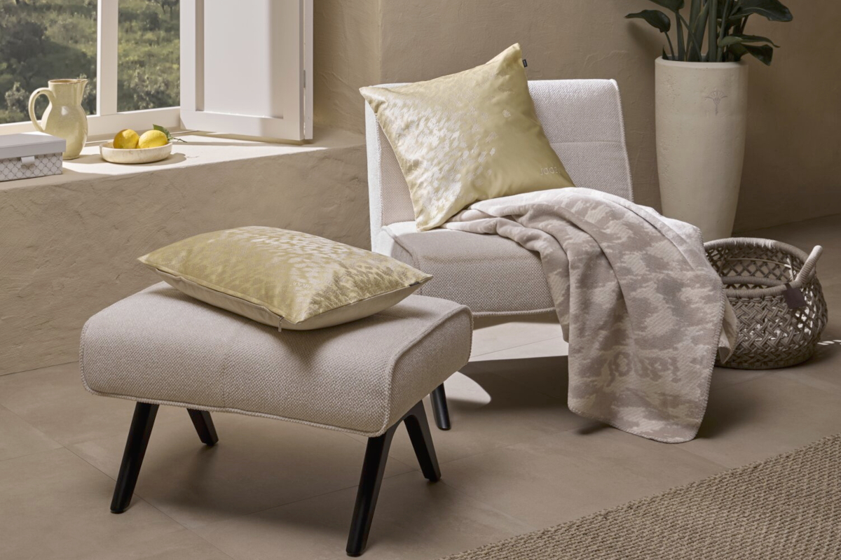 Ein helles, mediterranes Wohnzimmer mit Kissen der Serie Spirit in hellgelben Satin mit Animal Print-Design auf dem cremefarbenen Sessel Square und einem passendem Hocker