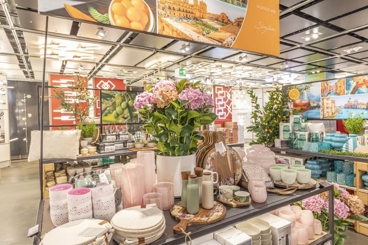 Ladenauslage mit farbenfrohen Küchenutensilien, rosa Hortensien und Dekoartikeln. Malerische Fotos und Grünpflanzen unterstreichen die lebendige Atmosphäre.