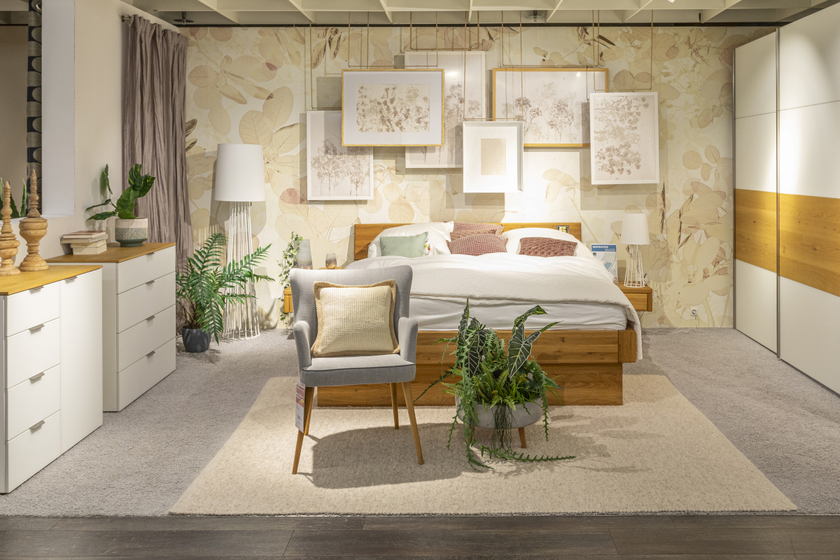 In einem Ausstellungsraum für Schlafzimmermöbel der Marke MAISON stehen das Massivholzbett Omaha mit integrierten Nachttischen und die Kommode Miami mit einer weißen Glasfront. Die Wand ist mit Bildern und einer Tapete mit zarten Pflanzenmotiven dekoriert.