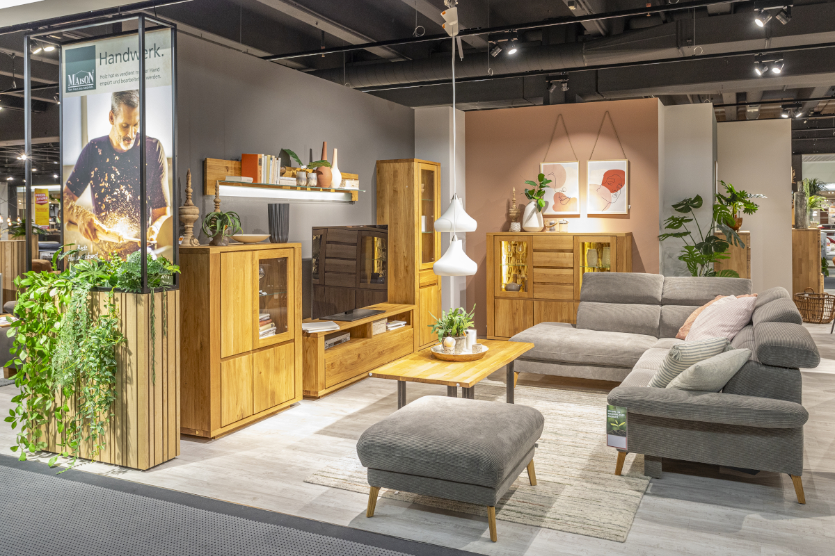 In einem Ausstellungsraum für Wohnzimmermöbel der Marke MAISON stehen das Sofa Arlington mit passendem Hocker in silbernem Cord und die vierteilige Wohnwand Boston aus Massivholz.