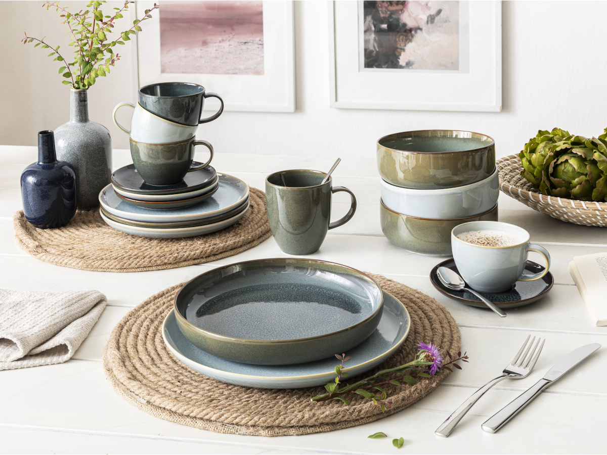 Ein weißer Holztisch mit Geschirr der Kollektion Crafted von Like. by Villeroy & Boch mit einer Effektglasur in in Blau- und Grüntönen.