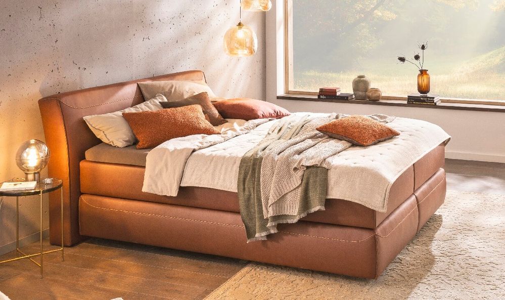 Gemütliches Schlafzimmer mit braunem Lederbett mit mehreren Decken und Kissen. Natürliches Licht strahlt durch ein großes Fenster mit landschaftlichem Ausblick.