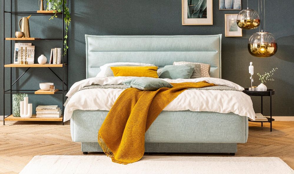 Modernes Schlafzimmer mit blaugrünem Bett, senffarbener Tagesdecke und Dekokissen. Ein Regal mit Büchern und Pflanzen, Pendelleuchten und gerahmter Kunst an der Wand.