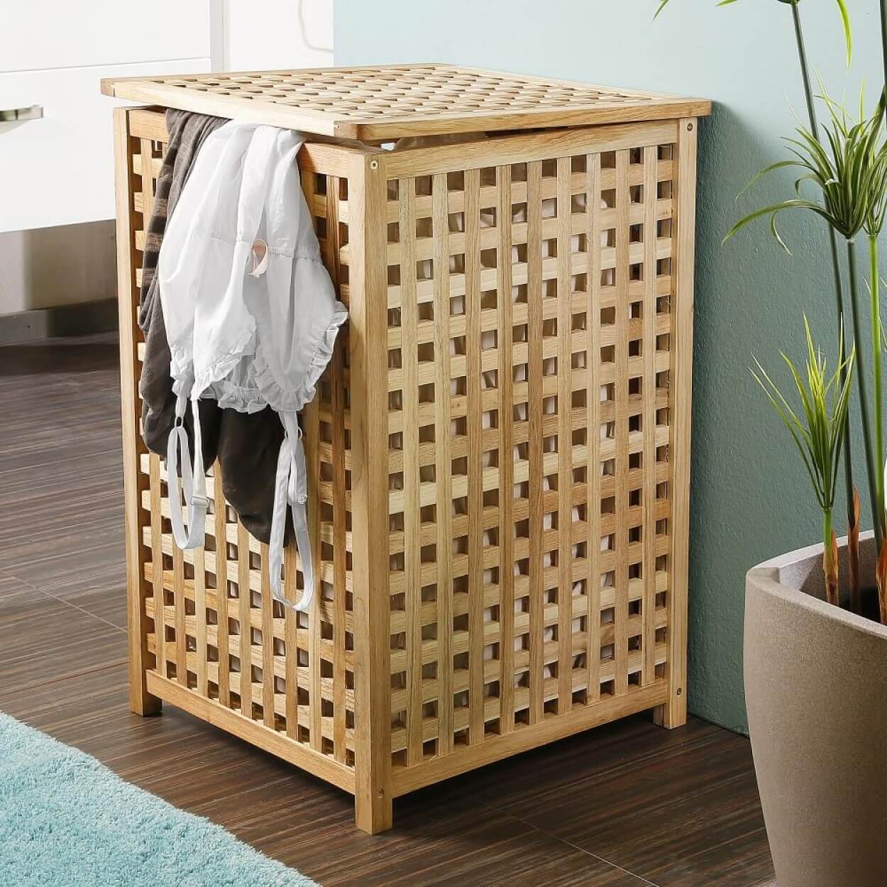 Holz-Wäschekorb tavia mit einem Gitterdesign, der Kleidung hält. Er steht auf einem gefliesten Boden nahe einer blühenden Pflanze und einer hellblauen Wand.