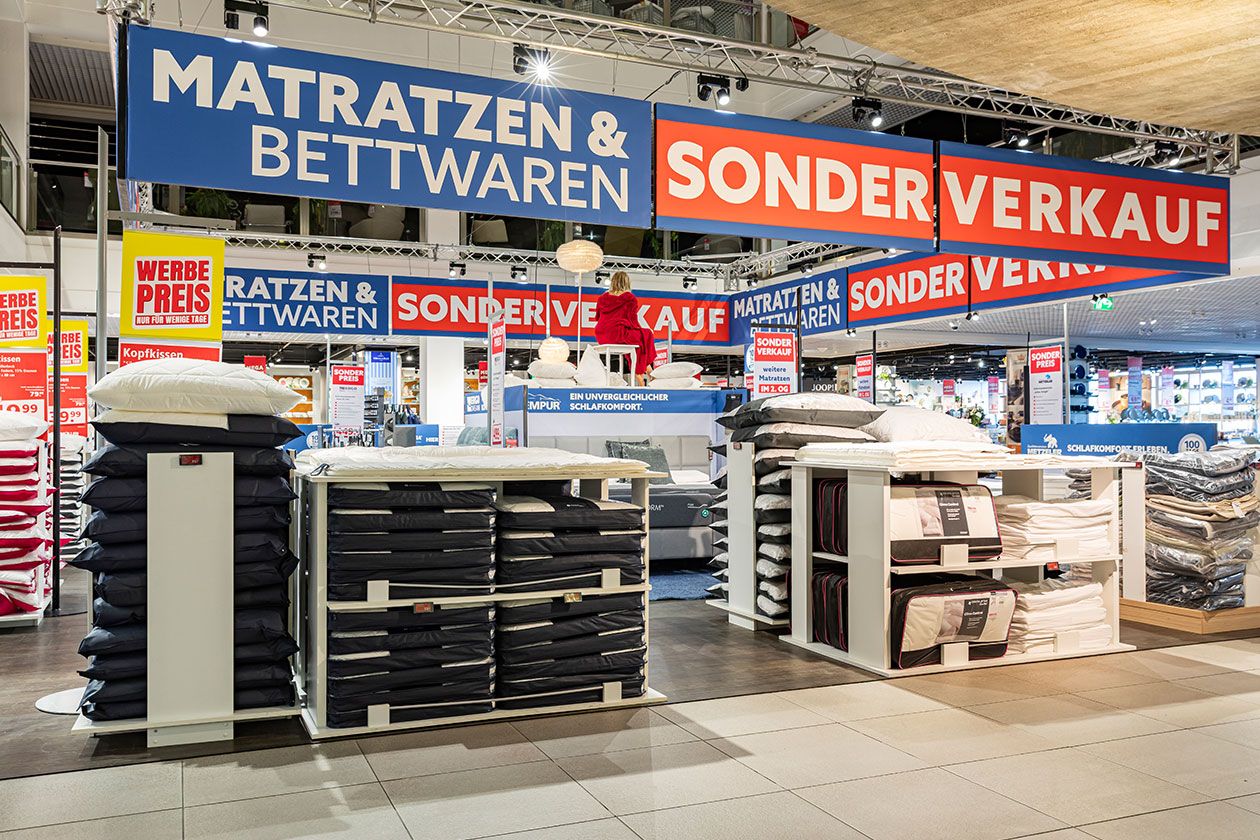 Im Lichthof eines SEGMÜLLER Einrichtungshauses werden Bettwäsche und Matratzen angeboten. Stapel von Kissen und Decken unter leuchtenden Schildern mit der Aufschrift „Sonderverkauf“.