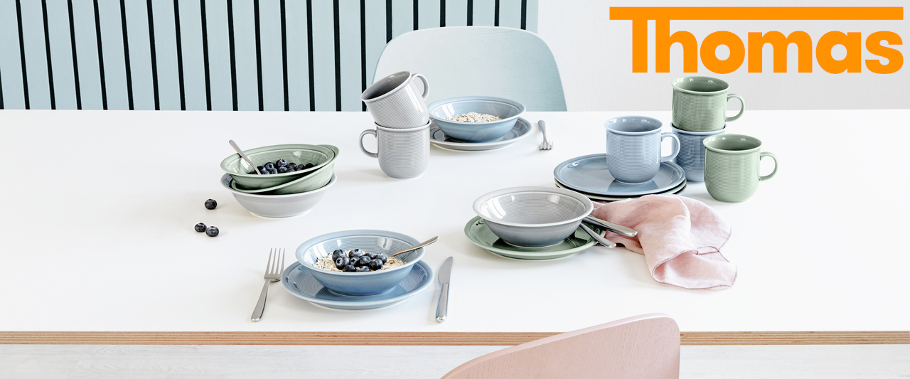 Auf einem weißen Tisch sind Schüsseln, Teller und Tassen der Serie Trend White & Colour der Marke Thomas aus Porzellan in den Farben Grau, Blau und Moss platziert.