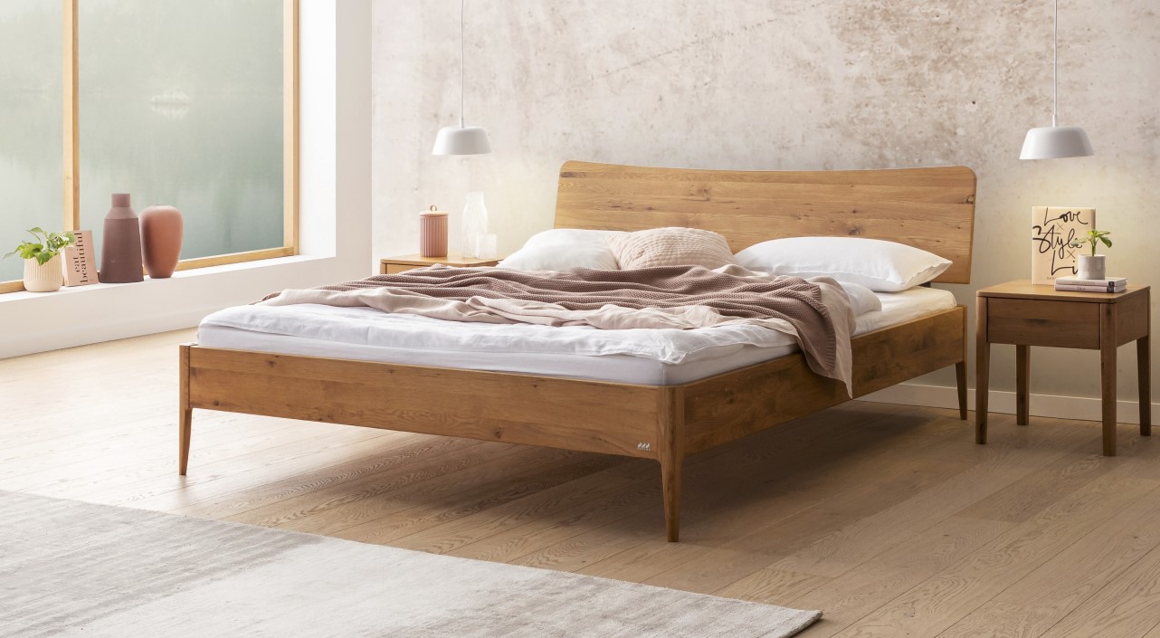 Minimalistisches Schlafzimmer mit einem Holzbett, weißer Bettwäsche und einer beigen Decke. Zwei Pendelleuchten und ein Fenster mit Milchglas runden die Szenerie ab.