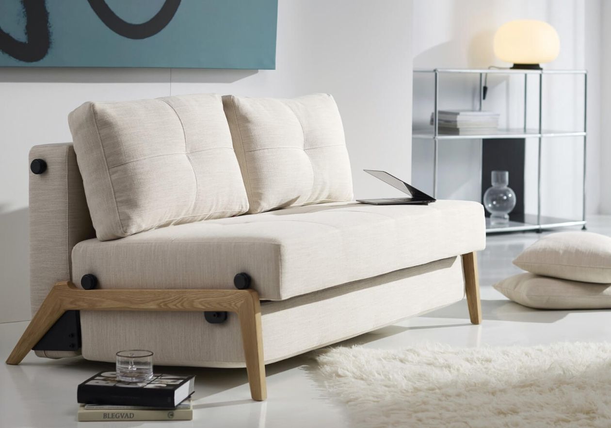 Modernes beigefarbenes Sofa mit Holzbeinen, zwei Kissen und einem Laptop darauf. In der Nähe ein Glasregal mit Dekoration und ein weicher Teppich auf dem Boden.