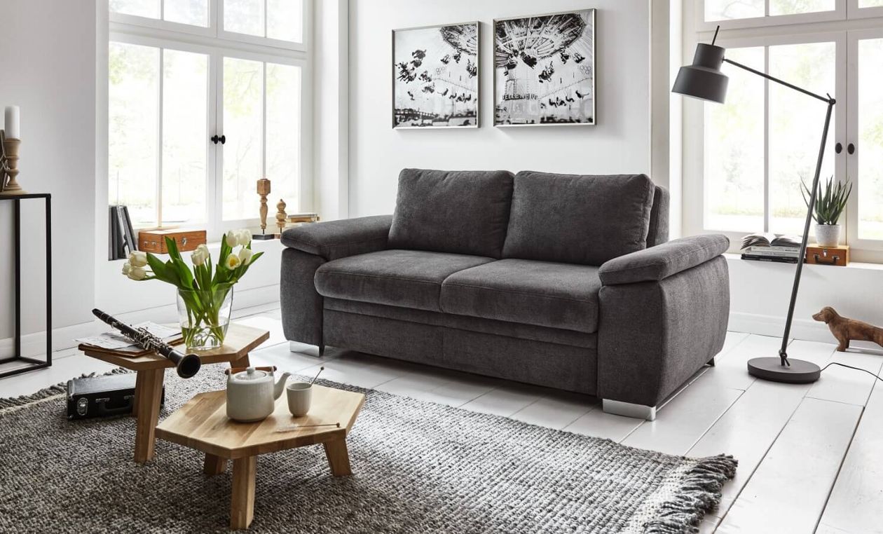 Moderne Wohnzimmereinrichtung mit einem grauen Sofa, einem Holztisch und einer Stehlampe. Wandkunst und große Fenster schaffen eine helle, gemütliche Atmosphäre.