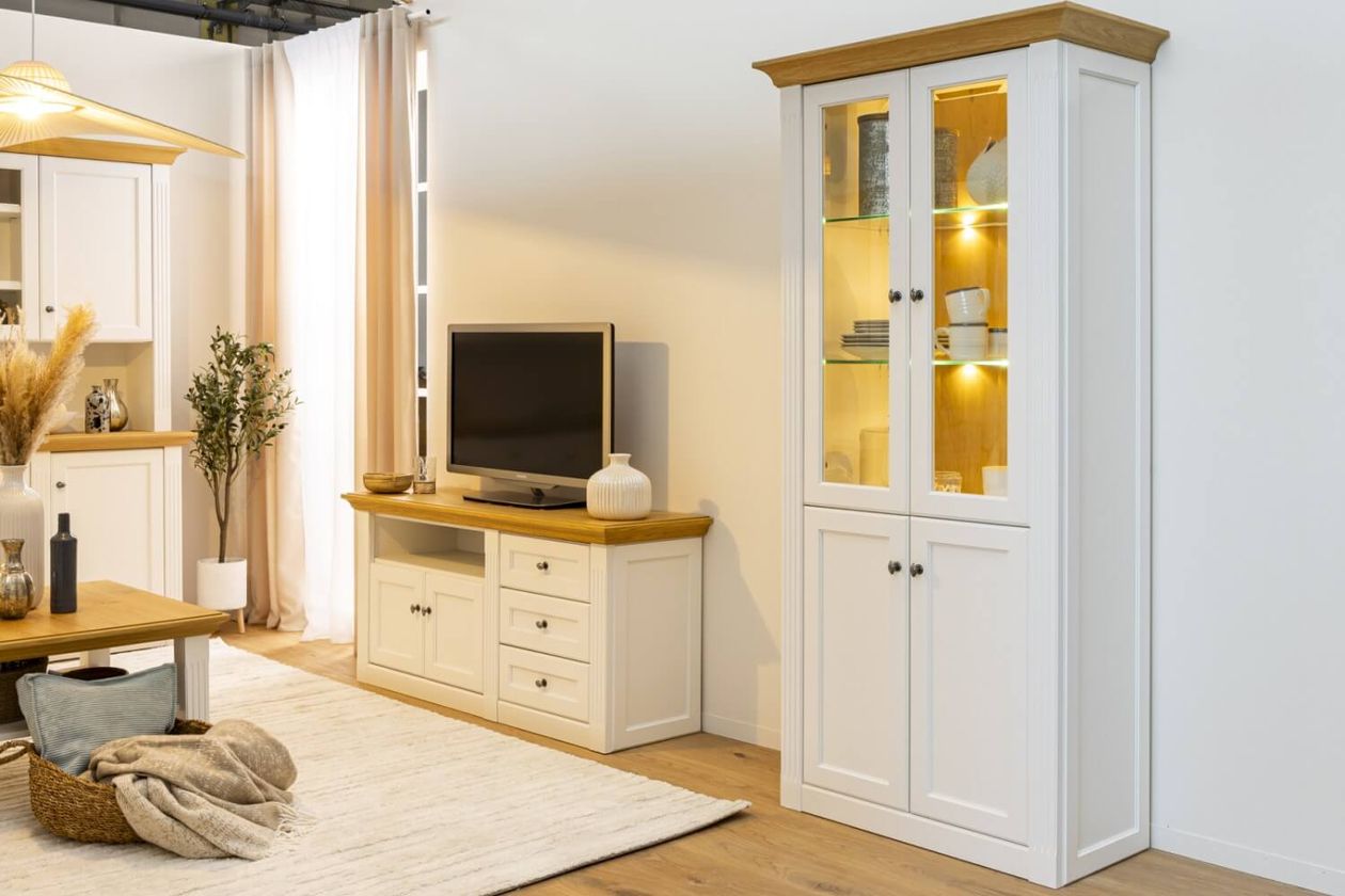 Ein gemütliches Wohnzimmer mit weißen Möbeln, einem Fernseher auf einem Holzlowboard und einem beleuchteten Vitrinenschrank.