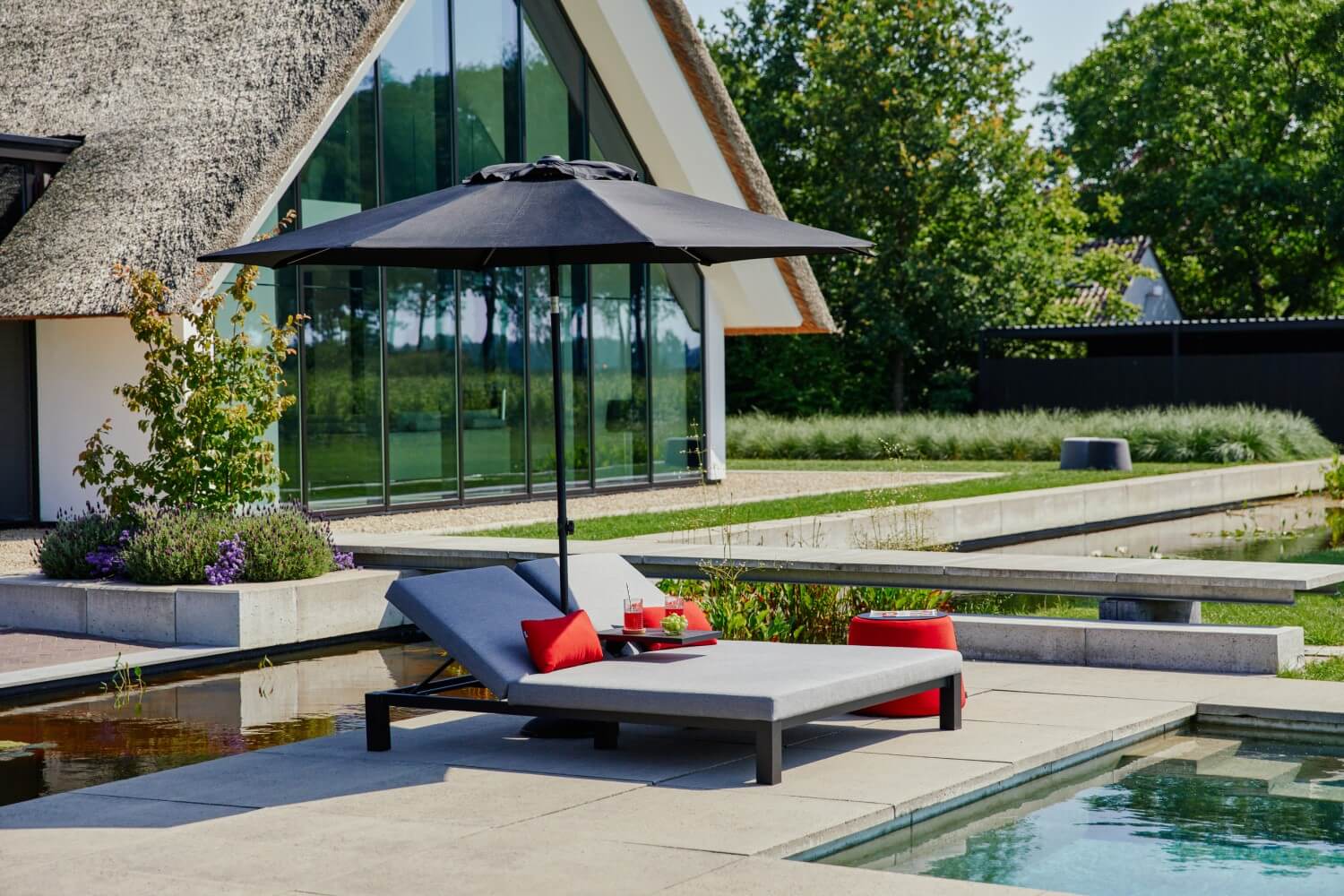 Moderne Poollandschaft mit einem eleganten Liegestuhl, roten Kissen und Sonnenschirm.