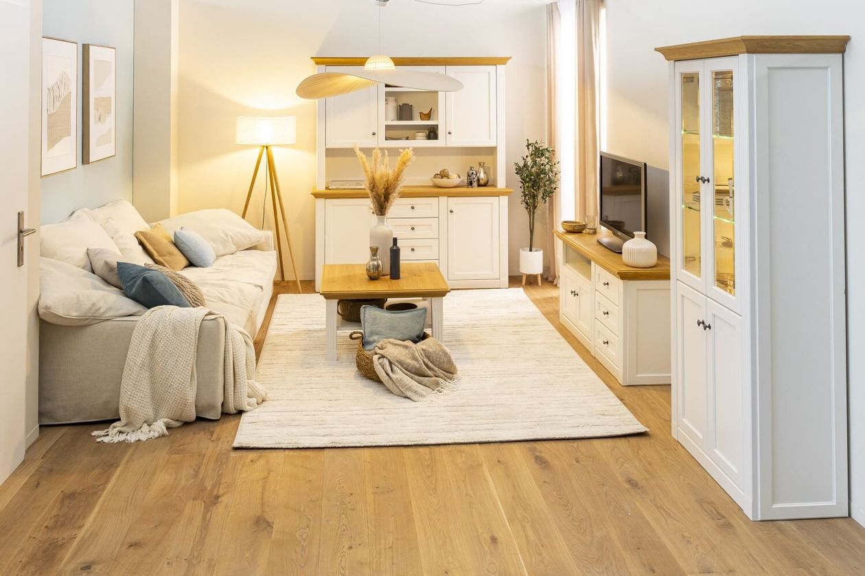 Gemütliches Wohnzimmer mit einem beigen Sofa, einem Holztisch, einem Fernseher und einem Schrank. Sanfte Beleuchtung und neutrale Dekoration schaffen eine warme Atmosphäre.