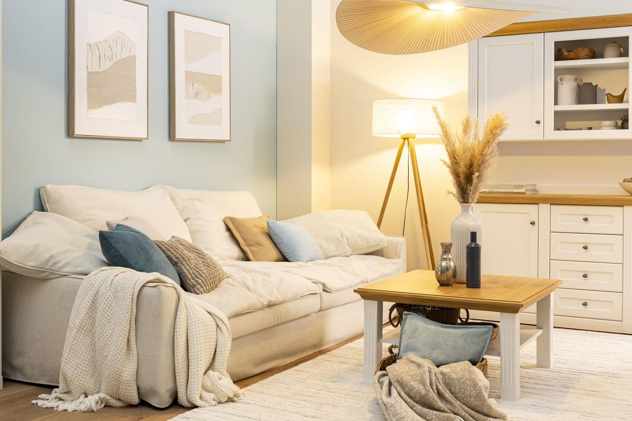 Gemütliches Wohnzimmer mit einem beigen Sofa, blauen und beigen Kissen, einem Holztisch und einer Stehlampe. Wandkunst und Schränke im Hintergrund.