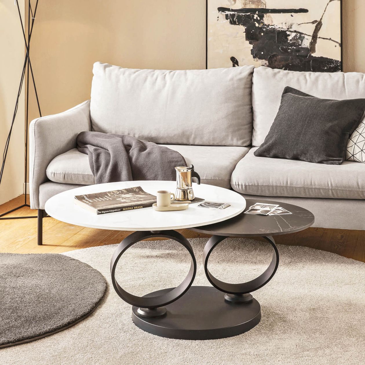 Ein modernes Wohnzimmer mit einem grauen Sofa, abstrakten Wandbildern und einem doppelten runden Couchtisch, auf dem ein Buch, eine Kaffeekanne und eine Tasse stehen.