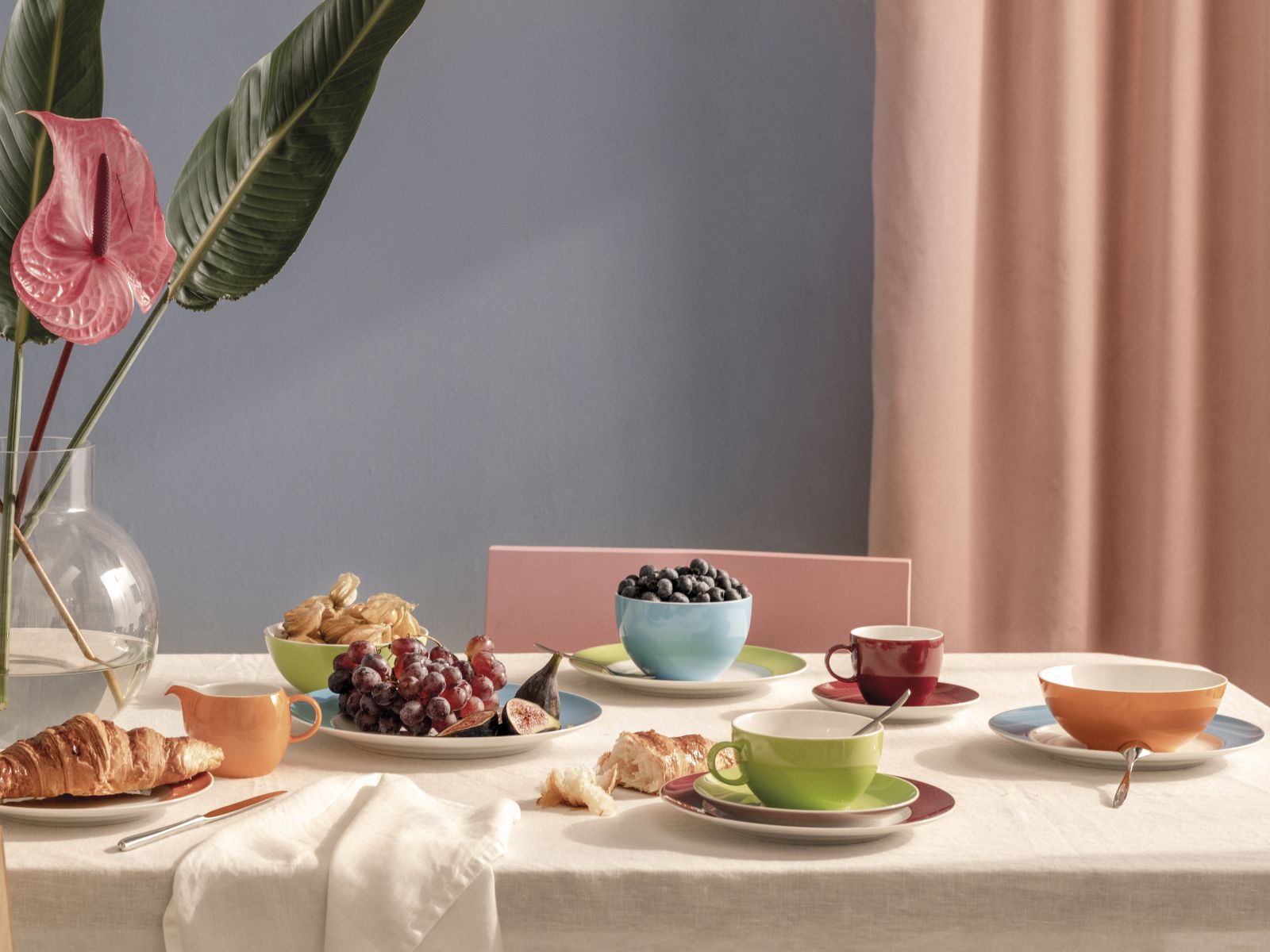 Auf einem mit Croissants und dunklen Trauben gedeckten Tisch stehen Schüsseln, Teller und Tassen der Serie Sunny Day aus dem Hause Thomas aus Porzellan in den Farben Rot, Orange, Grün und Blau.
