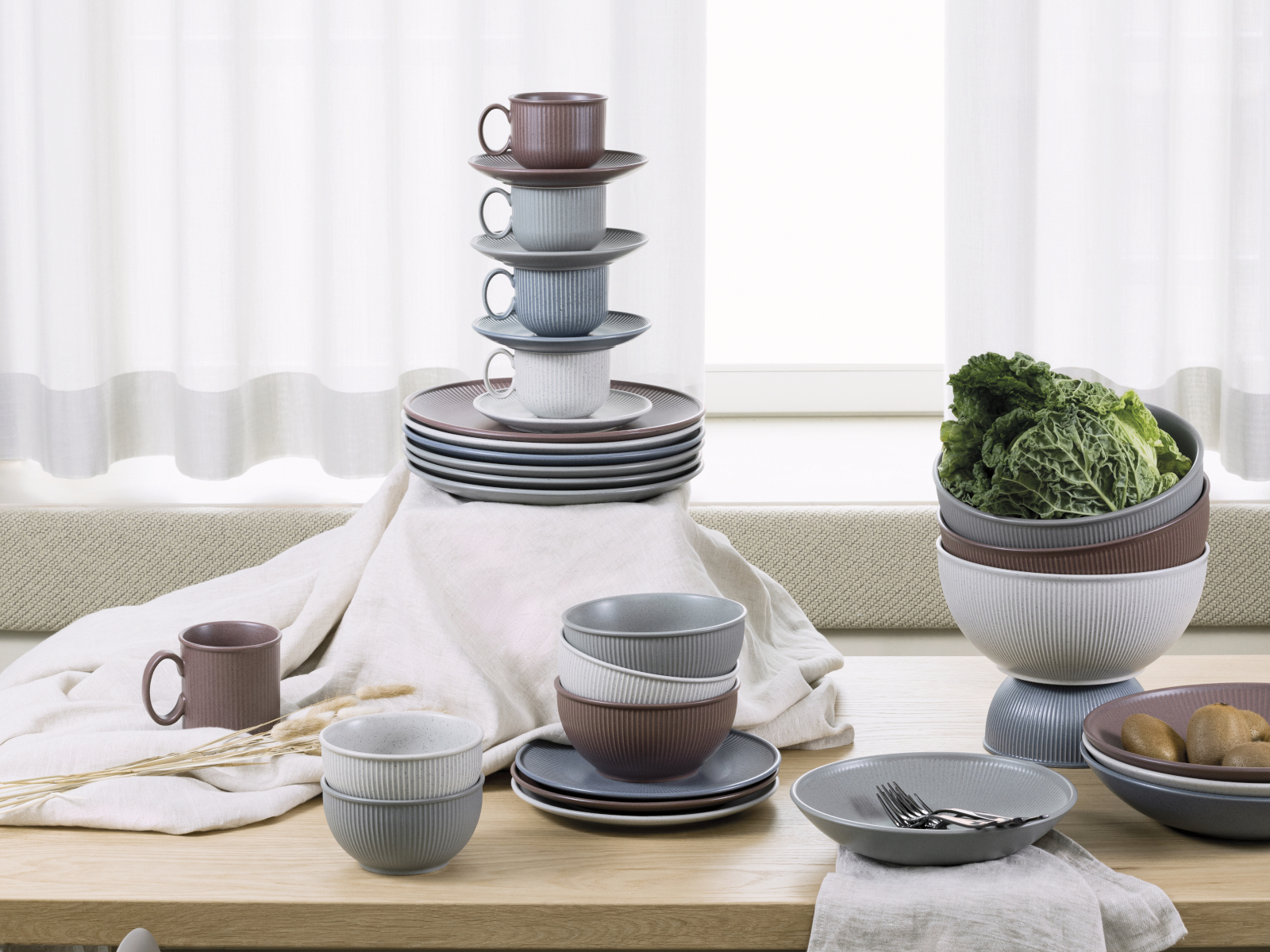 Auf einem Tisch sind Schüsseln, Teller und Tassen der Serie Clay aus blauem, braunen und grauen Steinzeug aus dem Hause Thomas dekorativ gestapelt.
