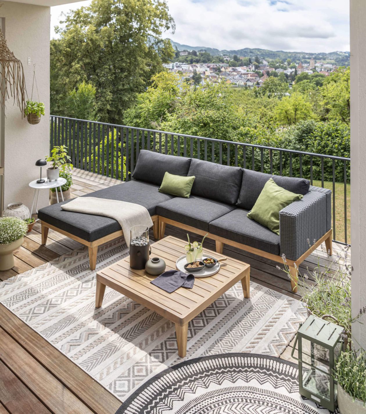 Ein stilvoller Balkon mit einer grauen Sofagarnitur, Holztisch und Pflanzen, mit Blick auf grüne Bäume und eine Stadt im Hintergrund.