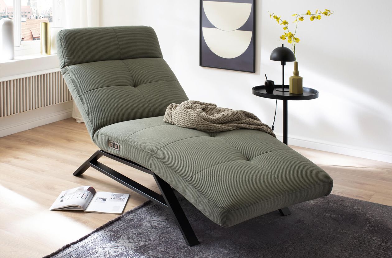 Grünes, modernes Liegesofa mit Kissen, daneben ein runder Tisch mit gelber Vase und Lampe, helle Fenster und grauer Teppich im Hintergrund.