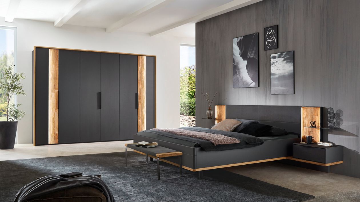 Modernes Schlafzimmer mit dunklem Bett, einem Schrank mit Holzakzenten und indirekter Beleuchtung, einer Sitzbank, Nachttischen und Wandbildern.