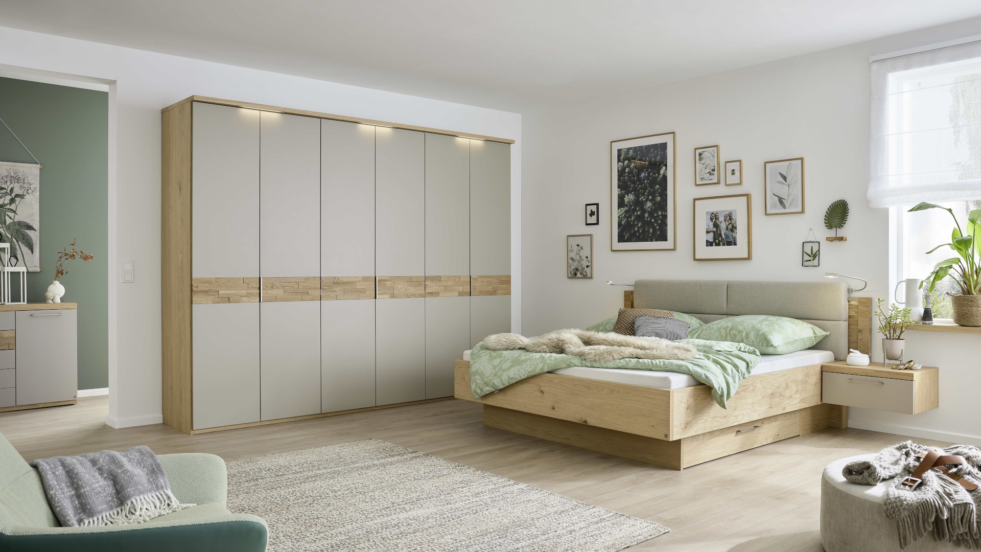 Die Schlafzimmerserie Minto 2.0 von Musterring mit einem Bett und Kleiderschrank mit Fronten in Lack in Taupe und Holzelementen aus Balkeneiche. Das Bett verfügt über ein gepolstertes, beiges Kopfteil.