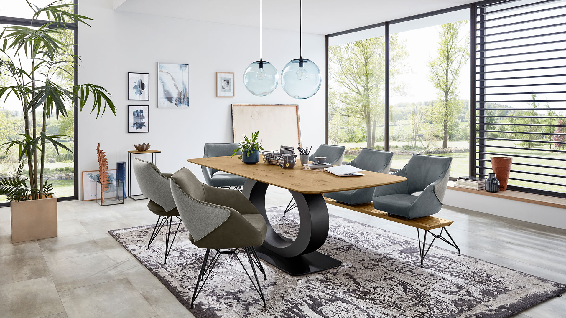 Ein modernes Speisezimmer mit Möbeln der Serie Brisko von Musterring. Zu sehen sind unter anderem der Esstisch Brisko, der dank seiner Platte aus furnierter Wildeiche und einem Gestell in auffälliger Kreisform auffällt sowie zwei Stühle Brisko SAEO mit braunem Lederbezug und grauem Stoff auf der Rückseite.
