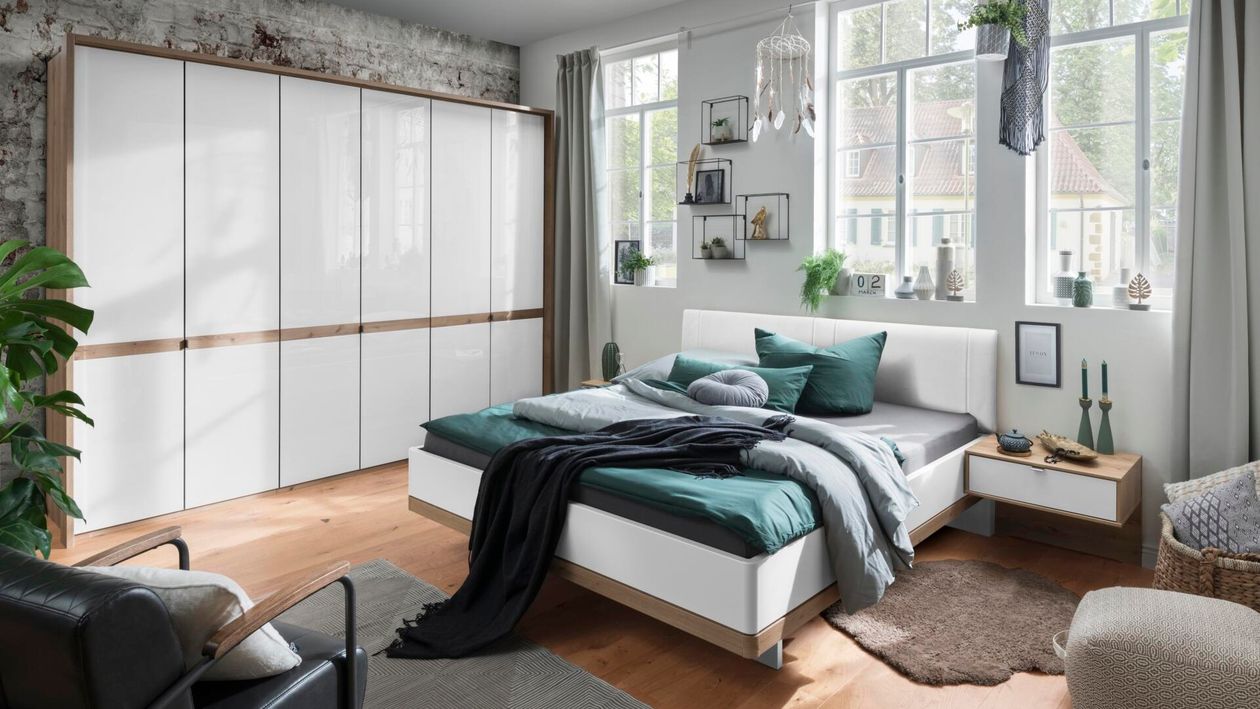 Das Schlafzimmer-Set Barcelona der Marke Wiemann mit einem Schrank mit weißer Glasfront und einem Bett mit Absetzungen in Bianco Eiche und einem Polsterkopfteil in Lederoptik.