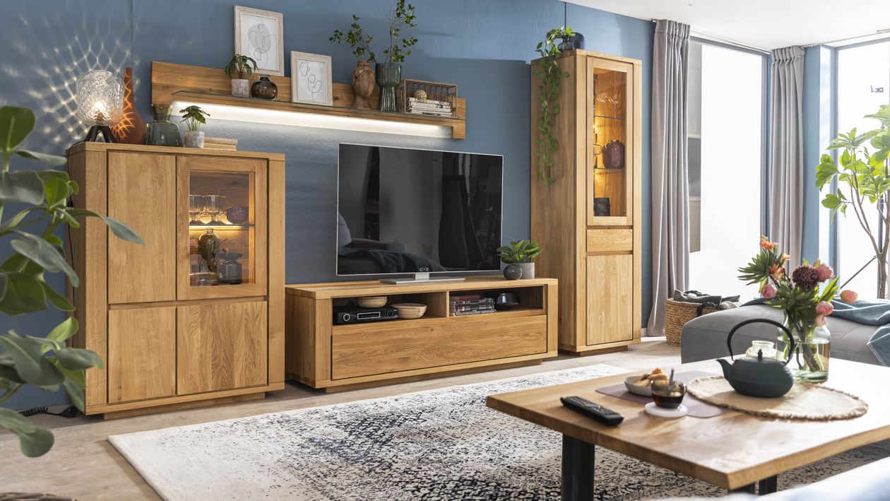 Modernes Wohnzimmer mit Holzmöbeln, großem Fernseher, Zierpflanzen und gemustertem Teppich. Blaue Wände und viel Tageslicht durch große Fenster.