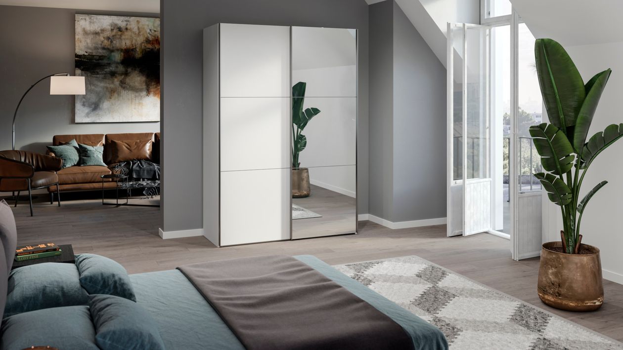 Weißer Schwebetürenschrank mit Spiegeltür neben einem Durchgang in ein Wohnzimmer mit Ledercouch. Klick führt zum Schrank im Shop.