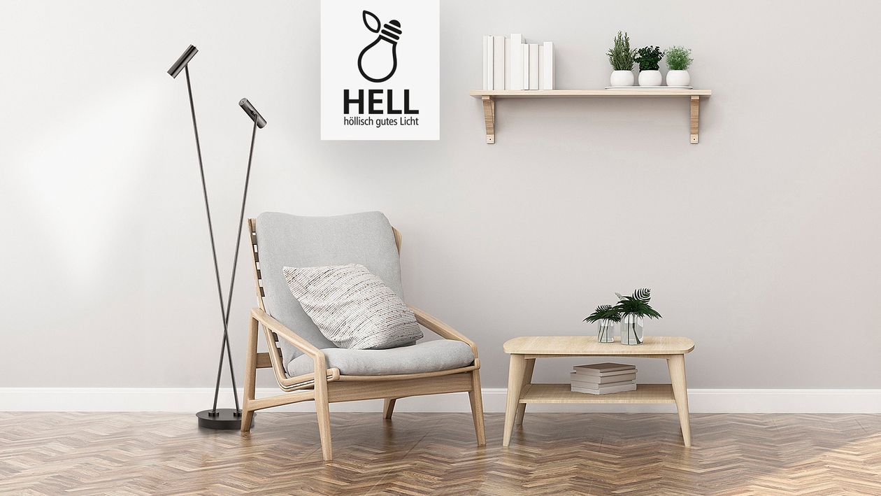 Eine schwarze Stehlampe der Marke Hell mit zwei Spots steht hinter einem Holzsessel mit grauen Kissen.